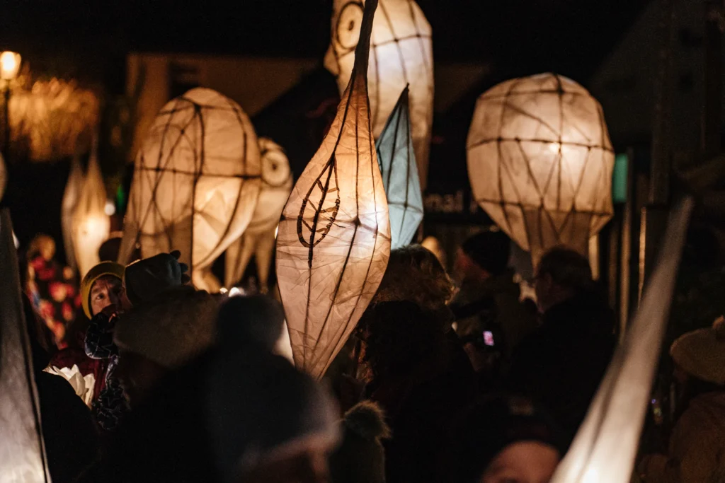 Close-up of glowing handmade lantern sculptures shaped like leaves and figures, carried by participants in a night parade.
