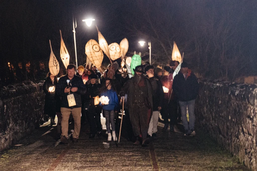 Group of people walking at night across a stone bridge, carrying lanterns and surrounded by glowing festival sculptures.