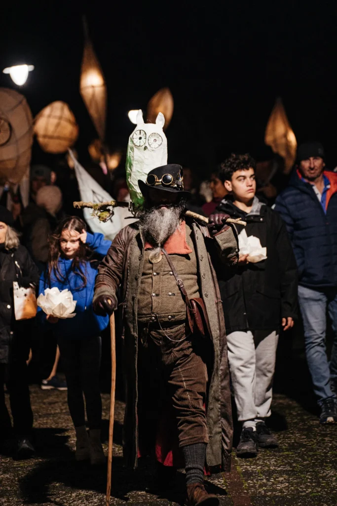Parade leader in costume walking at the front of a lantern procession, with families and glowing lanterns behind.
