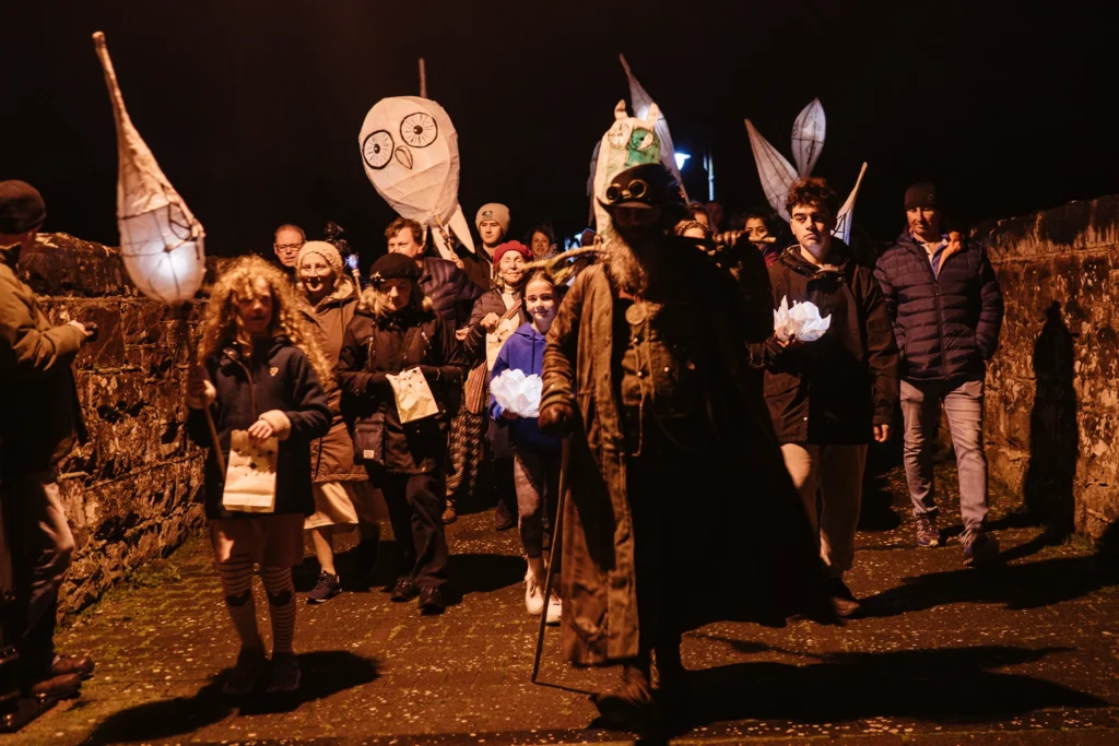Crowd following costumed performers at night, holding lanterns while large illuminated figures rise above the group.