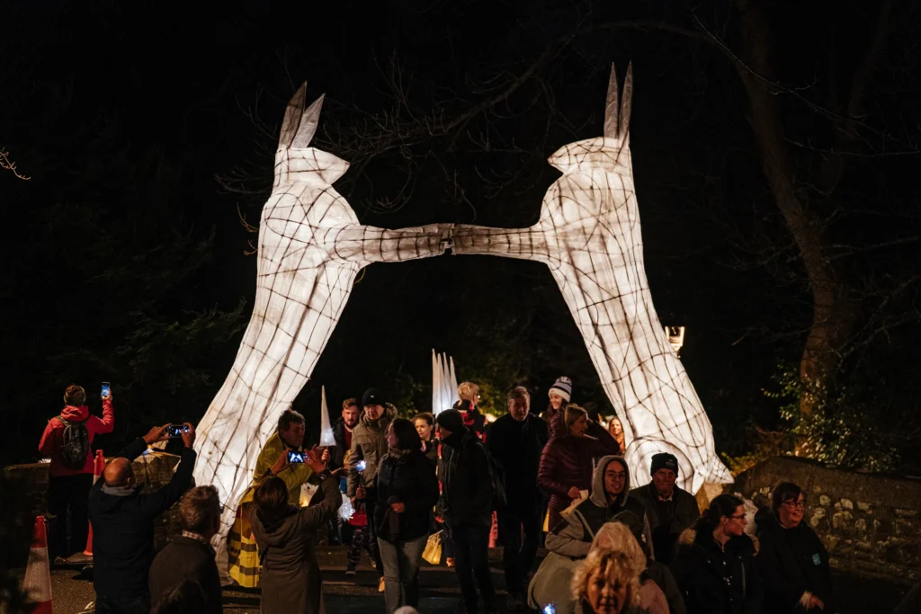 Large illuminated animal-shaped lantern sculpture towering above a crowd gathered at night in a wooded festival setting.