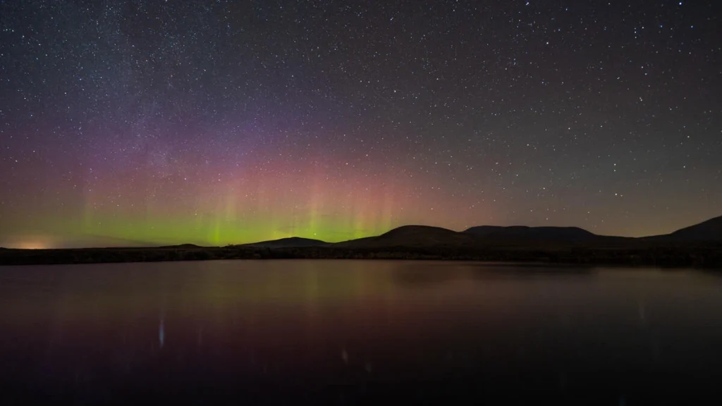 Aurora borealis above Mayo hills reflecting in serene lake, under starry night sky at Dark Sky Festival, Ireland.