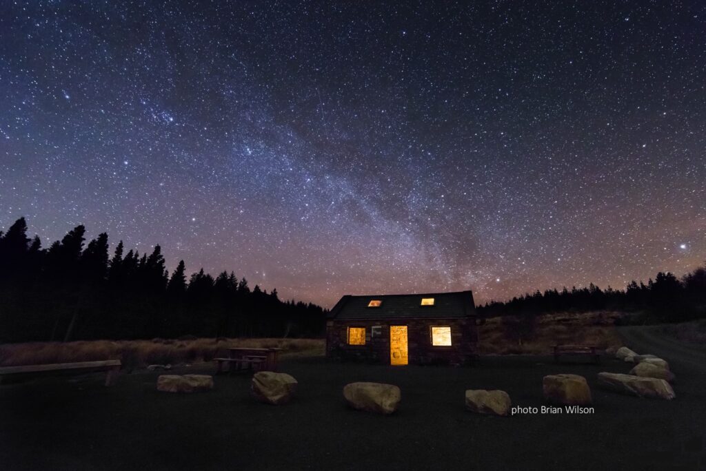 Starry night sky over a cosy, lit-up cabin amidst a dark forest setting. Perfect for stargazing and solitude.